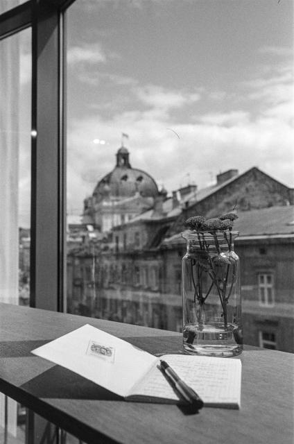 black-and-white film photo.
The view from the window of a cafe on the third floor. A large window covers the entire wall. There is a table near the window. There is a bouquet of flowers in a vase. Next to it is an open notebook with a pen.
The window offers a nice view of the old town street. The dome of the Lviv Opera House can be seen a little further away.