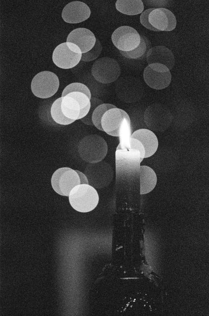 black-and-white film photo.
A Christmas candle is burning in the foreground. In the background you can see the lights from the garland on the Christmas tree. Lights are very blurred, so they look like large light bubbles
