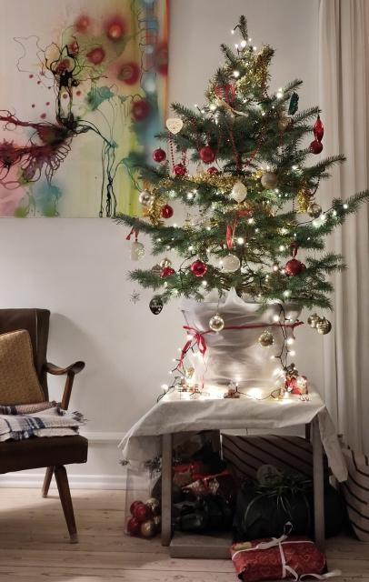 A Christmas tree in a pot, decorated with lights, tinsel and glass baubles stands on a low table with gift-wrapped presents underneath. A bright abstract painting is on the white wall behind 