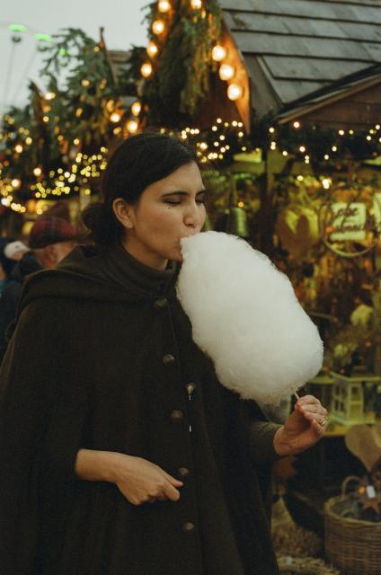 A beautiful woman with black hair, dressed in an olive-colored woolen coat with big brass buttons is holding white cotton candy in one of her hands and biting into it with determination. In the background, Christmas market stands with fairy lights and pine branches.