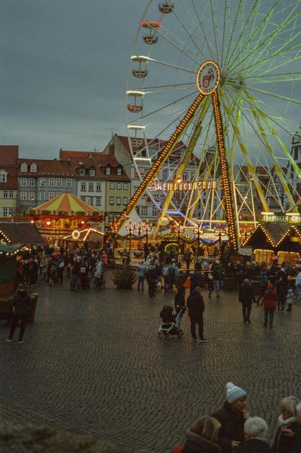 A crowd of people on a cobble stone plaza occupied by Christmas market stands and a ferris wheel, all decorated by fairy lights. There is no snow and the sky is grey as the sun is setting. 