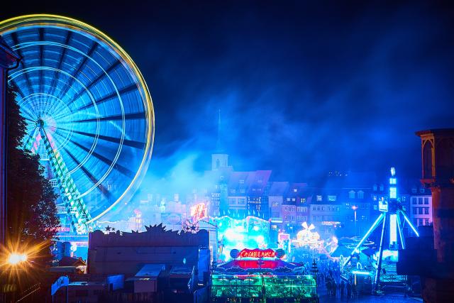 The Christmas market in Erfurt at night. On the left a blue lighted ferris wheel. On the right a Swing Coaster. A lot of little christmas stalls in the center. Behind this, a lot of houses frommthe city of Erfurt with a church tower in the dust.