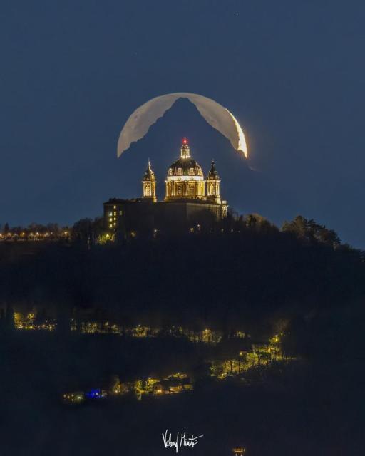 A tree-lined hill is shown topped by a majestic cathedral. Directly behind the cathedral is of a triangular-shaped mountain top. Directly behind the mountain is a crescent moon, although the exposure is long enough to see the rest of lunar circle.