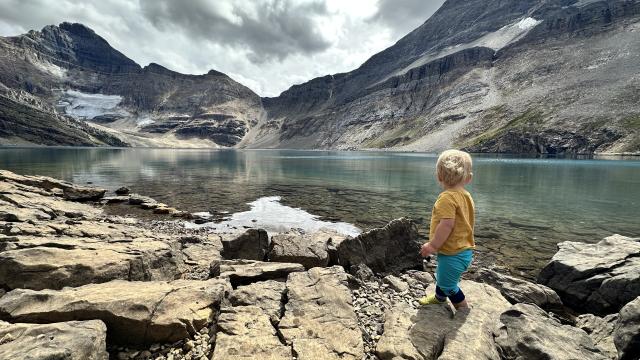 A young boy looks out across an alpine lake with rocky mountain peaks and a glacier in the background.