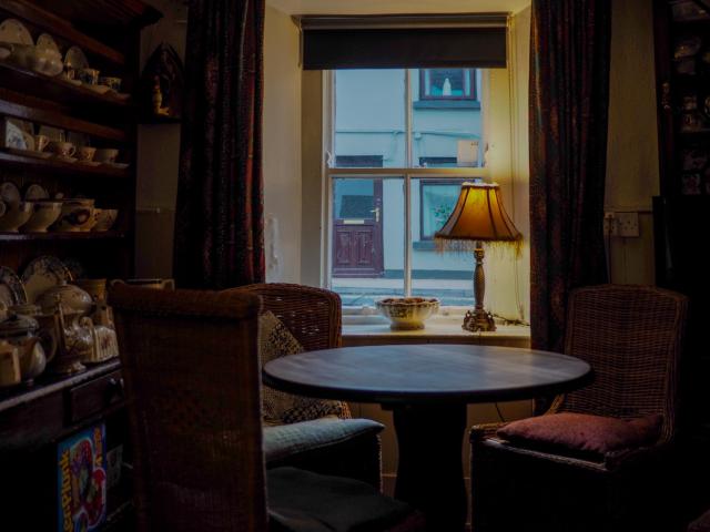 A photo of an empty, dimly lit corner of a bar with a round table and a few chairs by a window as seen front the inside. A table lamp with a yellow lamp shade and a small bowl are on the window ledge. The light on the table lamp is on. 