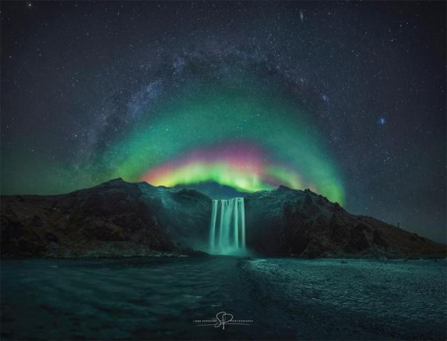 A waterfall is shown in the image center below a starry sky. Arching above the waterfall is a colorful aurora. Arching above the aurora is the central band of the Milky Way.