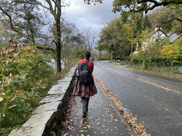 A woman in a red sweater and woolen plaid skirt is walking along an asphalt road covered in orange leaves under a grey sky, surrounded by yellowing trees on both sides, a body of water on the left and some houses on the right. 