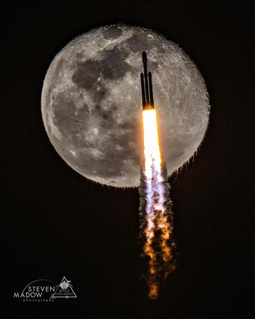 A rocket is pictured ascending during launch. A nearly full moon is behind it. The rocket exhaust, itself visible, causes the bottom of the Moon to appear unusually rippled.