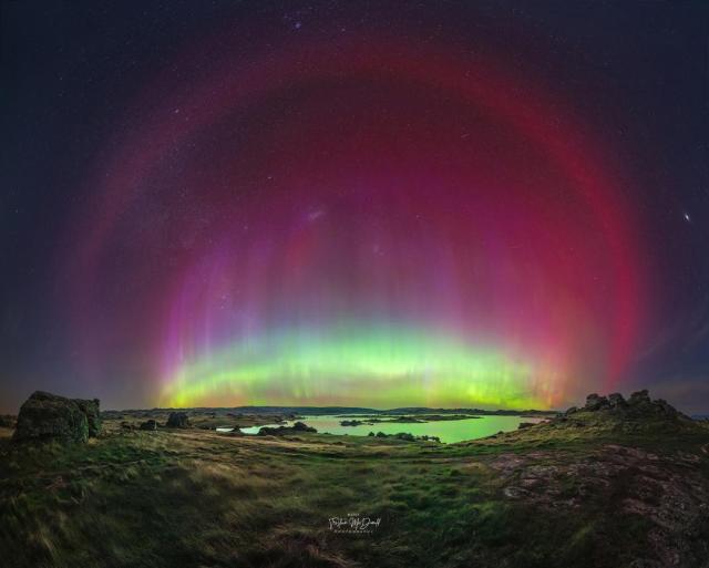 A flat landscape with a pond is imaged at night below a starfield. A multicolored aurora is seen in an arc across the image center. Around this arc is another red arc that is particularly smooth.