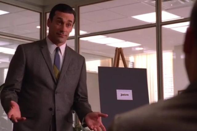 Don Draper from Mad Men in a suit stands in an office, facing another person, with an expectant expression and his hands outstretched.