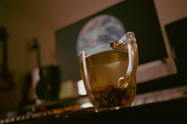 A glass mug with loose leaf tea, which has settled at the bottom.  In the background, out of focus, is a computer monitor with a picture of Earth from space.