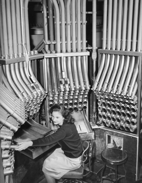 Woman routing canisters among approx. 200 pneumatic tubes.  According to "Growing Up in CHicago" on Fabebook, this is "1947 - Helen Serros routes money through tubes in the Marshall Fields Pneumatic Tube Room, 111 North State Street, Chicago, Illinois. The tubes average 90 seconds in a cash sale transaction."