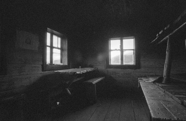 a black-and-white film photo.
Interior of a wooden shepherds' house in the mountains.
In the room in the left corner there is a wooden table with benches.
A bunk bed can be seen on the right.
The sun shines through two windows into the room.