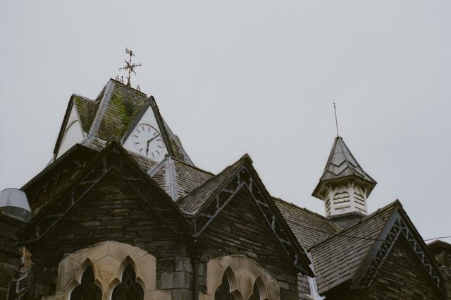 A roof on a grey stone building featuring a clock tower, a wind vane, a second tiny tower, and loads of moss, under a grey sky. 