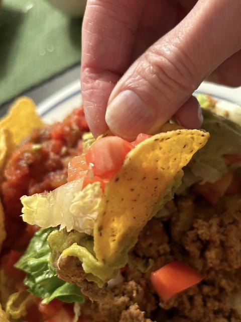 my thumb and forefinger holding a round corn tortilla chip that is perfectly bent like a taco shell, and is full of taco meat, lettuce, tomato, salsa, and guacamole 