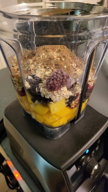 blender with all ingredients. Varied mix of frozen dark berries, frozen mango, + frozen spinach, oats, water, linseed, maple syrup.