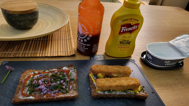 a sandwich open as 2 halves on a blue chopping board which rests on a wooden table. 2 toasted wholemeal rye breads, with creamy spread, scissorcut chives +flower. Twin spirals of yellow French's squeezy mustard, and red Sriracha extra chilli sauce, the bottles visible behind. 2 Air fried veggy fingers complete the sandwich, ready to be closed up.