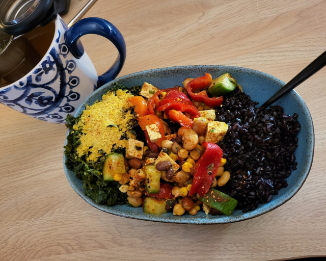 A ceramic boat dish holds a portion of black rice, steamed kale with a little lemon juice and Noosh flakes and a good helping of the batch salad made up thread. A black fork rests alongside the food in the dish. A cup of tea stands next to it on the desk.