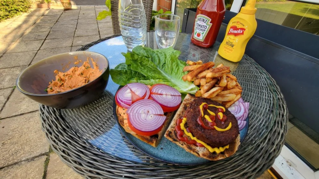 A blue ceramic plate of open faced burger with a fries stack, lettuce leaves and extra trimmings alongside. A black and blue bowl holds a red spicy hummus with chives for dipping fries (chips). Behind on the glass topped outdoor table sits water glass & squeezies of heinz ketchup and french's mustard.