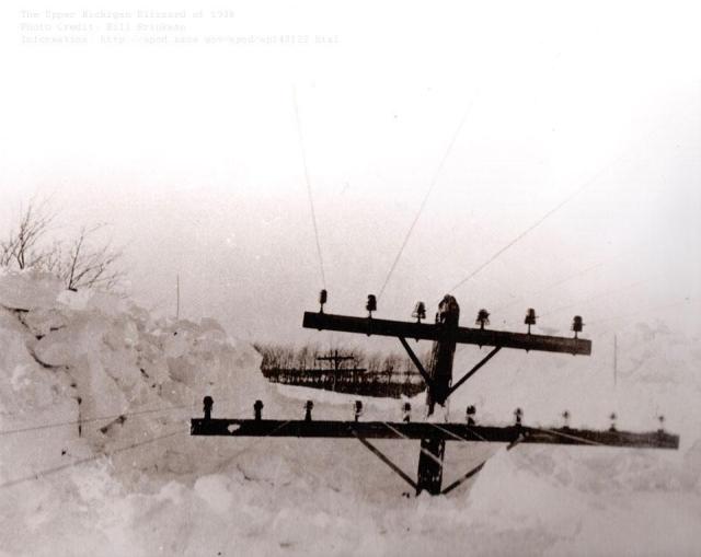 A telephone poll is shown surrounded by snow. In the background, another telephone poll is visible, as are some distant trees.