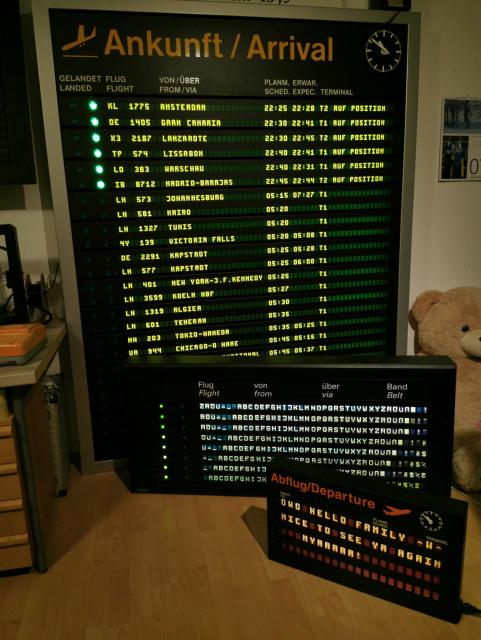 Three airport information displays in varying sizes. In the back is a very tall (around two meters) board with 24 lines showing actual arrivals at my local airport. On the floor, in front of it, is a smaller board (around 60 cm tall) with 8 lines, showing the alphabet as a test text. Slightly to the right and in front of that is yet another, smaller one, about 40 cm tall, with 4 lines, showing "OWO HELLO FAMILY ^W^ NICE TO SEE YA AGAIN NYAAAAA!" 