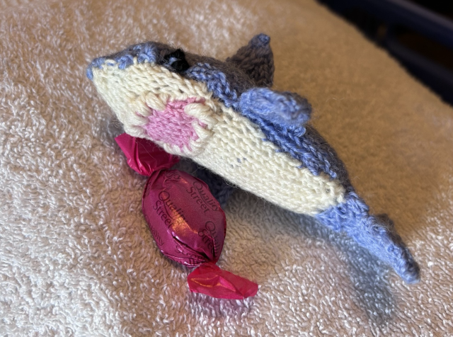 Small knitted blue shark holding a pink wrapped sweetie