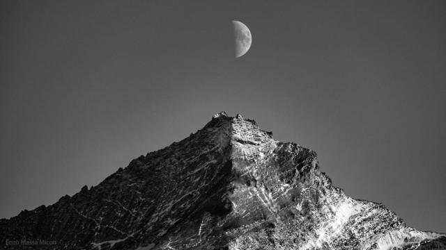 A black and white image of the Moon and a mountain are shown. Both are half lit by the Sun, with the other half shadowed. The half-moon is directly above the mountain peak.