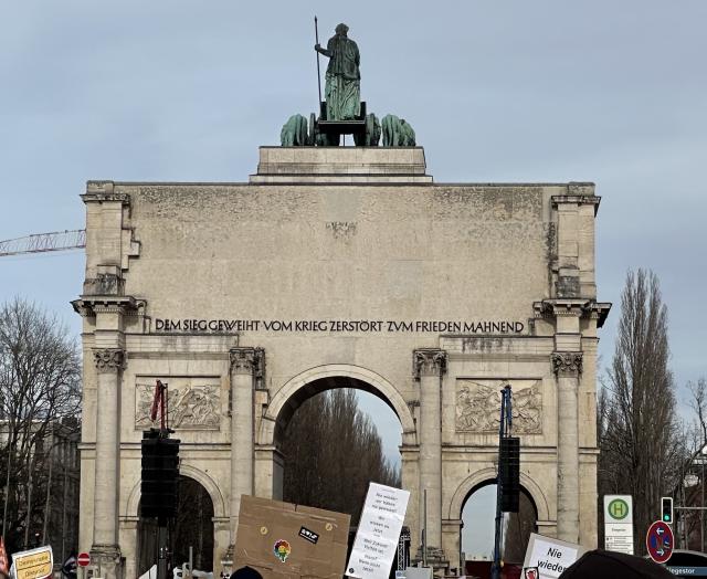 Triumphbogen mit einer Statue auf der Spitze und Inschriften, umgeben von Schildern einer Demonstration, mit einem Kran im Hintergrund links.
Inschrift: „Dem Sieg geweiht - Vom Krieg zerstört - Zum Frieden mahnend“