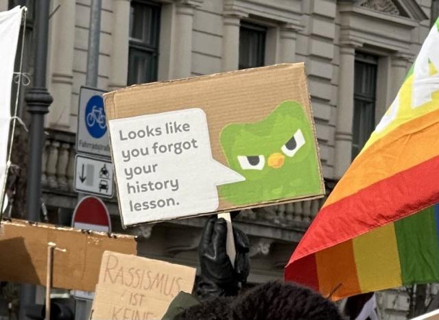 Ein Protestschild mit einer grünen Eule und der Aufschrift "Sieht aus, als hättet ihr euren Geschichtsunterricht vergessen" sowie Teile anderer Schilder und eine Regenbogenflagge.