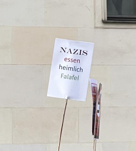 Ein an einem Stock befestigtes Schild mit der Aufschrift "NAZIS essen heimlich Falafel" vor einem Gebäudehintergrund.