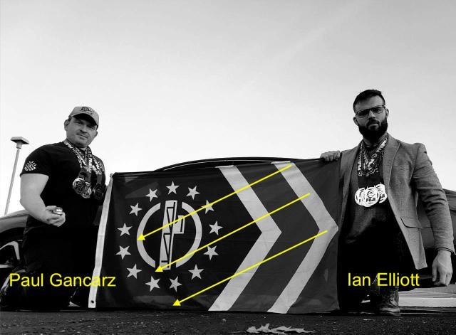Black & White photo of Paul Gancarz and Ian Elliott at the FUJI Allentown Winter Championships. Paul is on the left and Ian is on the Right. They're squatting down in the parking lot of the venue, holding a Patriot Front flag. They both wearing medals with FUJI on them.
