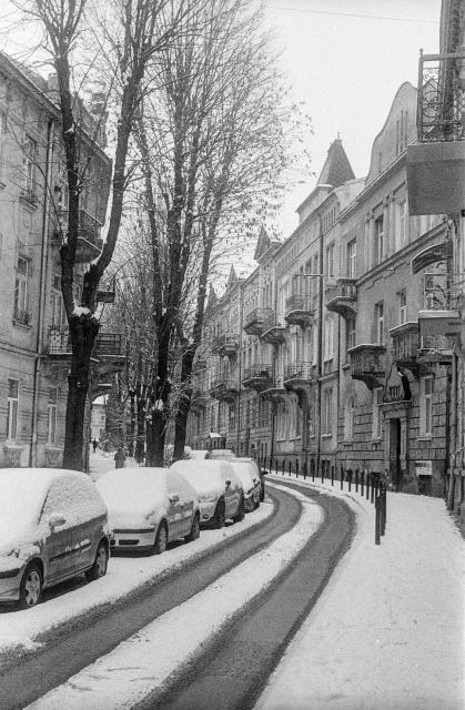 a black-and-white film photo.
A small street in the old town. Three-story houses stretch on both sides. The street turns gently to the left.
On the left side are parked cars covered with snow. The sidewalk on the right is also covered with snow. The road in the middle has two lanes of ruts in the snow from cars