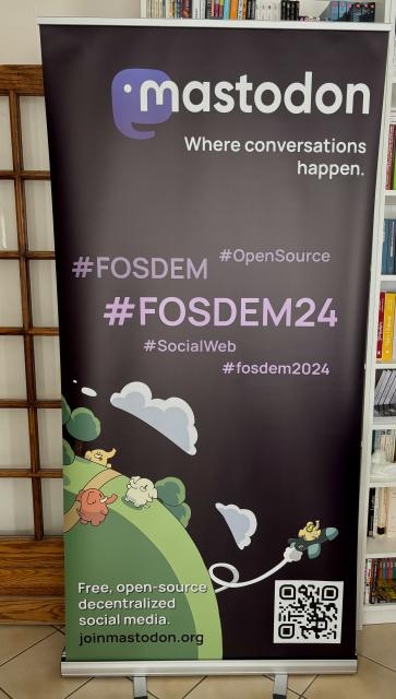 A roll-up banner with the Mastodon logo, the tagline "Where conversations happen", a cloud of hashtags #FOSDEM, #FOSDEM24, #OpenSource, #SocialWeb, and #fosdem2024, an illustration of colorful elephants on a green landscape and some clouds, a QR code, the second tagline "Free, open-source decentralized social media" and the address joinmastodon.org.