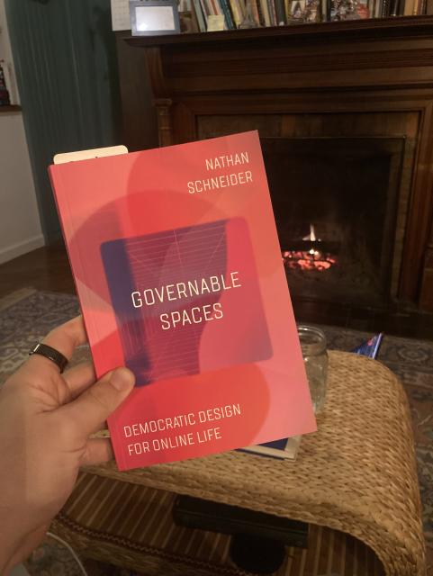 A photo of a book called Governable Spaces by Nathan Schneider. It's red, in a living room, with a fire behind. 
