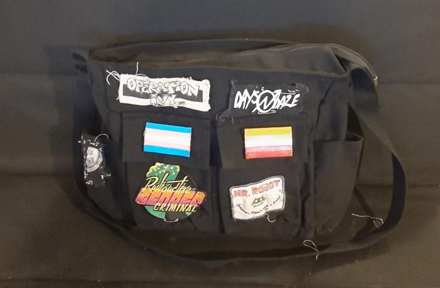 the black canvas messenger bag i've been sewing stuff onto. it has trans and lesbian flags and mr. robot and radioactive gender criminal and op ivy and apes of the state and days n daze patches