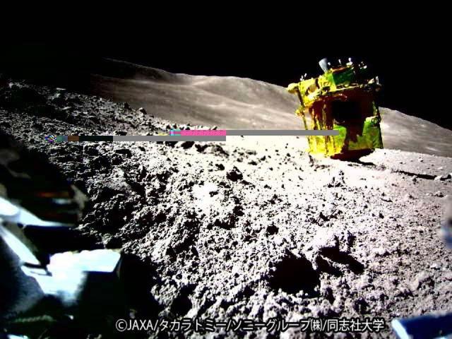 The lunar surface is shown with a box-like gold-colored machine in the middle. A close inspection of the machine reveals that its thrusters are at the top, so it is on its side. The background sky is dark. Two horizontal lines are an artifact of the digital imaging and not part of the lunar landscape.