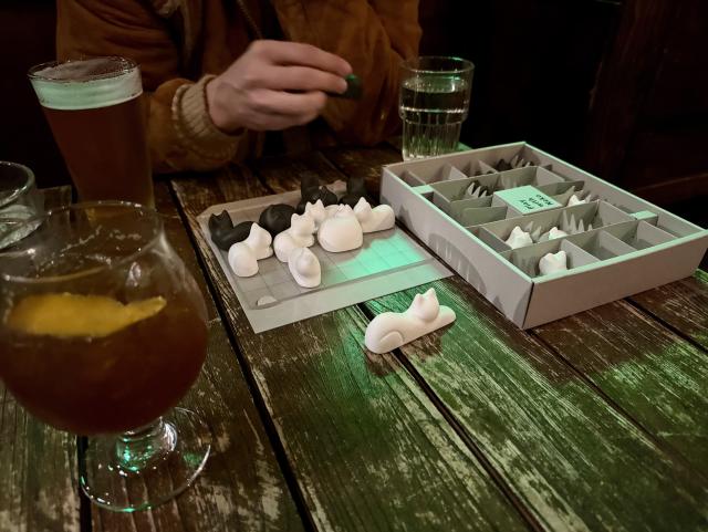 A shot of the game from further away. There are a couple of cocktails on the table and a hand is about to place a black cat on the board. 