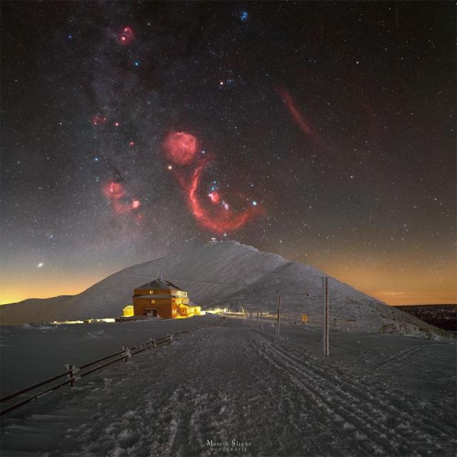 A snowy landscape is pictured with a big hill in the center. Above the hill is a starfield with the stars and nebulae of the constellation Orion appearing, with the red glow of the nebulas in great contrast to the dark sky and bright snow.