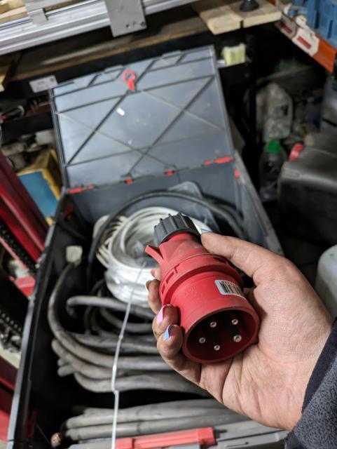 photo of my hand holding an iso/iec 3p+n+pe 16a plug. there's a huge crate of *thicc* cables in the background.