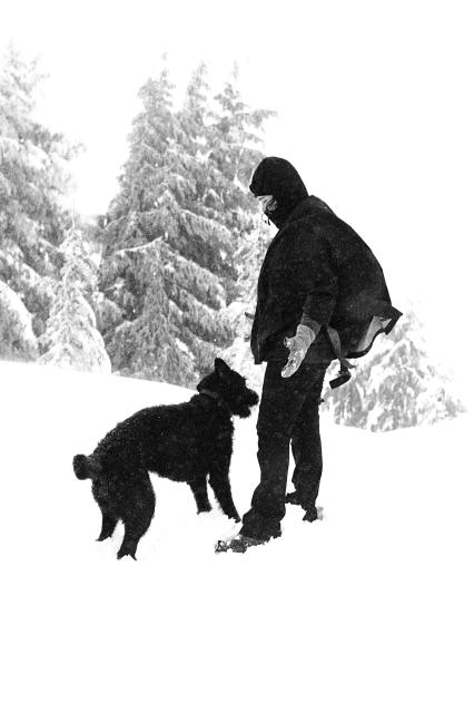 Person in winter clothing standing in snow with a dog, with snow-covered trees in the background.