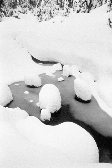 Black and white scene of a snow-covered landscape with a small stream meandering through it, featuring rounded shapes of rocks or boulders blanketed with thick snow.