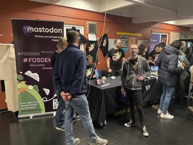 A table full of Mastodon merchandise, a roll-up with the Mastodon logo, and a bunch of people wearing Mastodon t-shirts in a hallway. 
