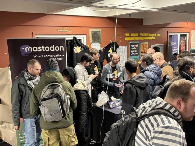 Lots of people talking to one another across the Mastodon table at FOSDEM 2024. There is a dark purple pull-up banner on the left with the Mastodon word mark and the phrase “where conversations happen” underneath it in white text  - conversations happened!