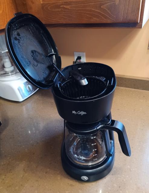 A black Mr. Coffee coffeemaker on a kitchen counter. The water reservoir flap is up and hinges to the left. The only access for pouring is from the right. 