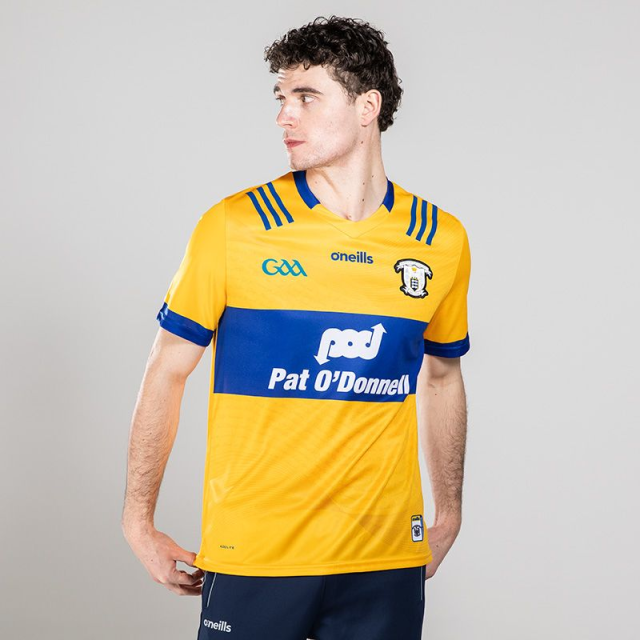 Male white model wears a hurling top of An Clár, it is gold with blue highlights and features a large advert for Pat O'Donnell.