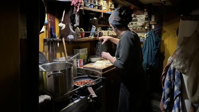 Person making dinner in a ship gallery with a tall pot and fiddles on the shelves. 