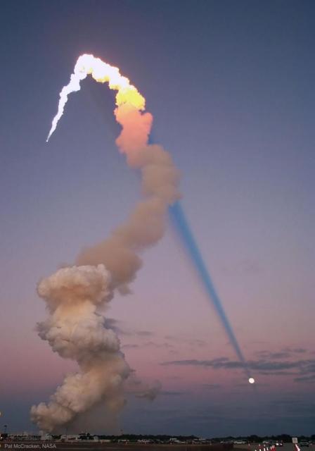 The long plume of a launching rocket is seen on the left side of the image. The upper part of the plume is bright, while the lower part is smokey brown. The bright part of the plume is illuminated by the Sun and casts a long and dark shadow corridor across the image. The shadow appears to end on a Full Moon.