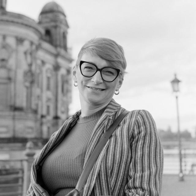 b/w analog square portrait of a middle aged woman with light skin and short light hair, smirking, wearing glasses with thick black frames and a striped jacket