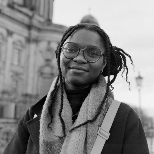 b/w analog square portrait of a young Black woman with black dreads and large glasses, wearing a fuzzy scarf and dark wool coat