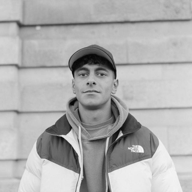 b/w analog square portrait of a young man with medium-dark skin and curly black hair. wearing a baseball hat, hoodie, and an outdoor jacket